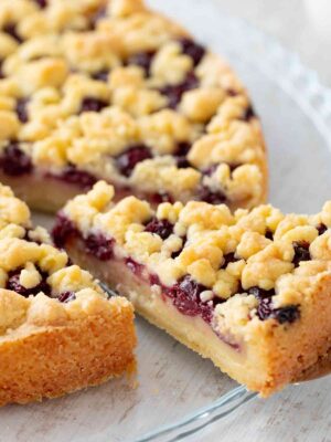 Nahaufnahme eines veganen Kirsch-Streuselkuchens mit einer Schicht Pudding auf einem gläsernen Kuchenteller. Ein Stück wurde herausgeschnitten.