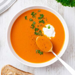 Tomatensuppe mit Kokosmilch auf einem Teller, garniert mit einem Klecks Kokosmilch und frischer Petersilie. Ein Löffel mit Suppe liegt im Teller.