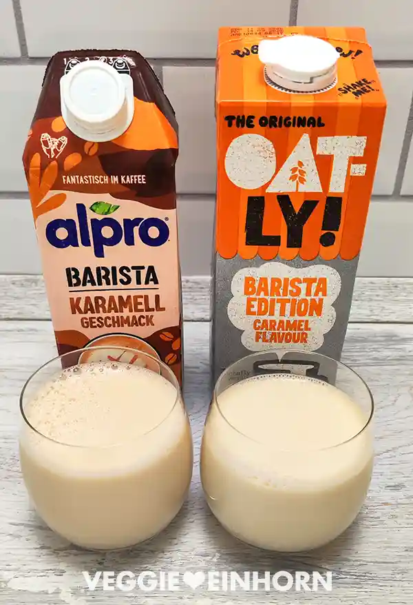 Zwei Gläser mit heller karamellfarbener Barista-Milch stehen auf einem weißen Holztisch. Dahinter stehen zwei Getränkepackungen, links Alpro Barista Karamell Geschmack, rechts Oatly Barista Edition Caramel Flavour.