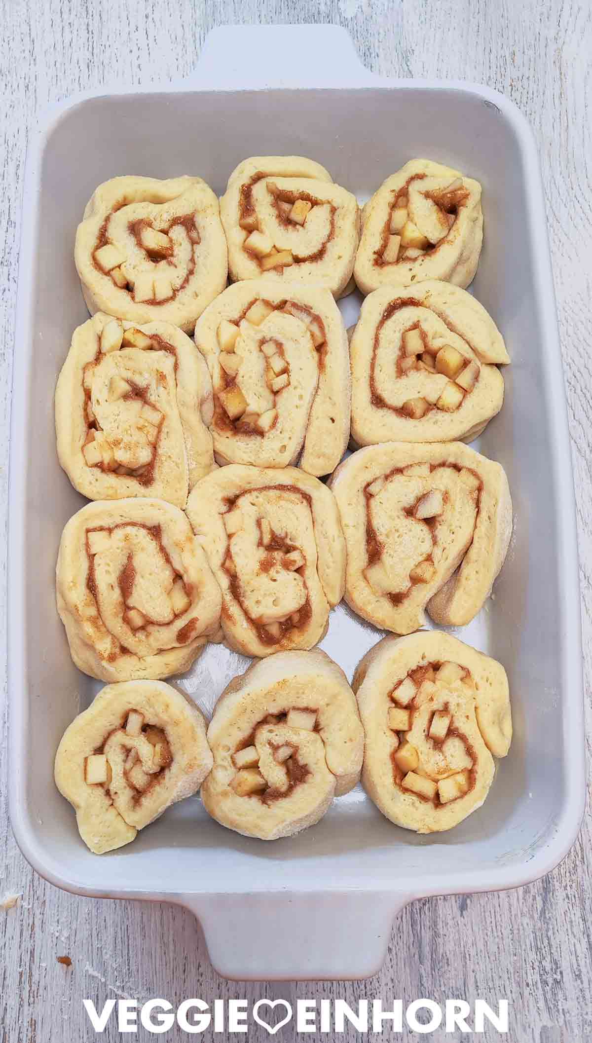 Zwölf ungebackene Apfel-Zimt-Schnecken liegen in einer weißen Auflaufform.
