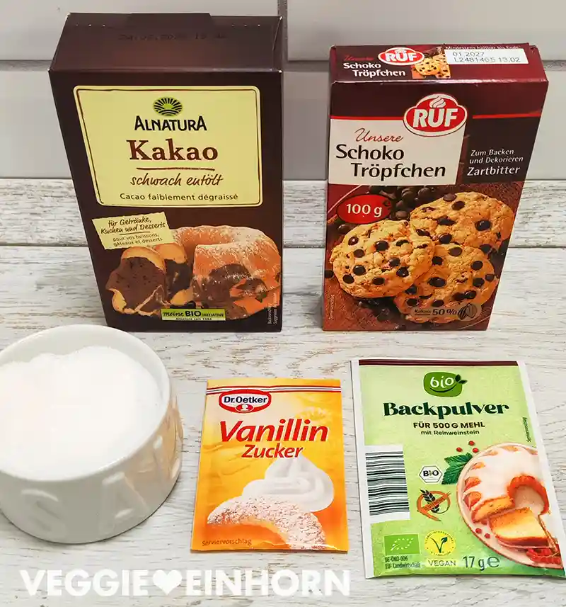 Mehrere Backzutaten wie Kakaopulver, vegane Schokotropfen, Vanillinzucker, Backpulver und Salz stehen auf einer hellen Holzoberfläche bereit.