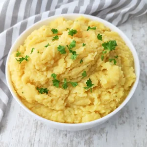 Veganes Kartoffelpüree in einer weißen Schüssel auf einem hellen Holztisch, dekoriert mit frischer Petersilie und einem grau gestreiften Küchentuch im Hintergrund.