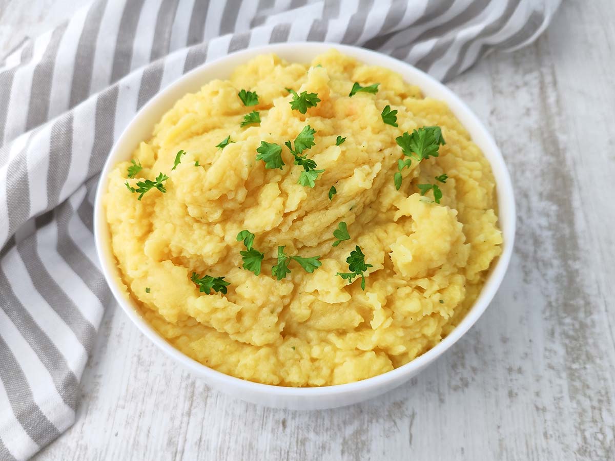 Weiße Schüssel mit cremigem veganem Kartoffelpüree auf hellem Untergrund, garniert mit Petersilie und neben einem grau gestreiften Küchentuch.