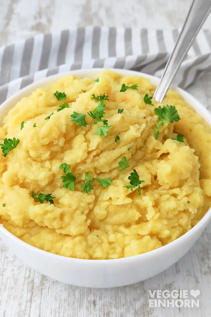 Bestes veganes Kartoffelpüree Nahaufnahme von veganem Kartoffelpüree in einer weißen Schüssel, mit Petersilie bestreut und einem Löffel im Püree.