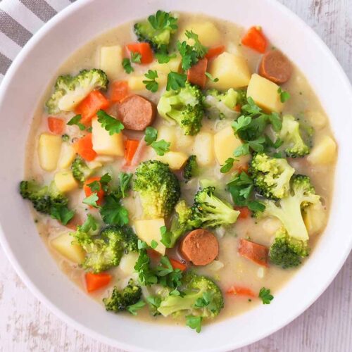 Veganer Gemüseeintopf mit Brokkoli, Kartoffeln, Möhren und veganen Würstchen, serviert in einem weißen Teller.