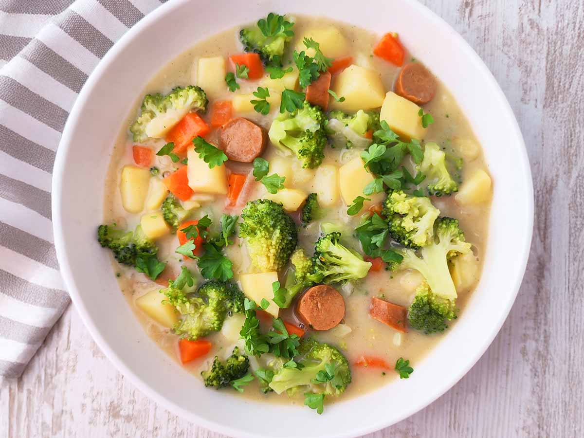 Veganer Gemüseeintopf mit Brokkoli, Kartoffeln, Möhren und veganen Würstchen, serviert in einem weißen Teller.