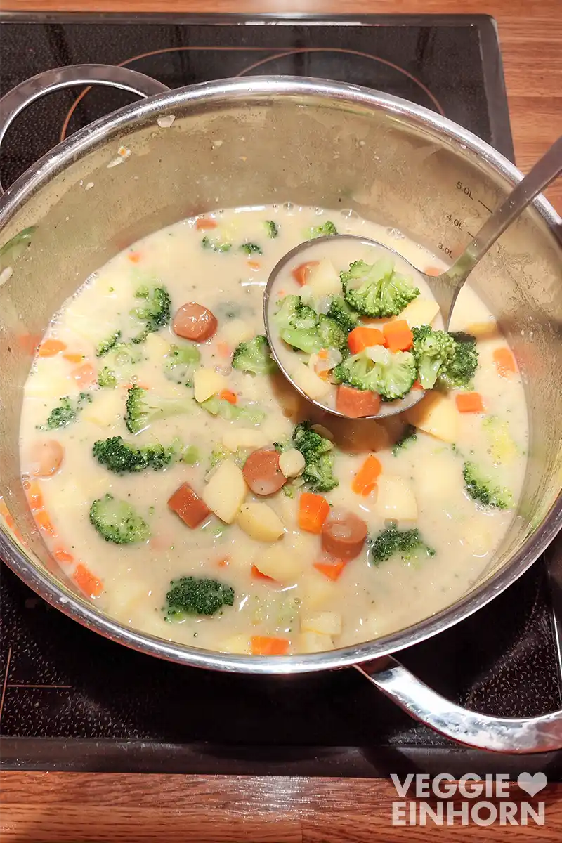 Ein Topf mit veganem Gemüseeintopf auf dem Herd, darin Brokkoli, Kartoffeln, Möhren und vegane Wiener Würstchen. Eine Kelle schöpft Eintopf heraus.