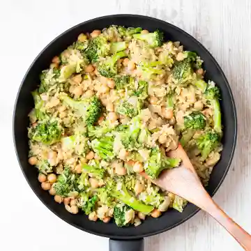 Vegane Brokkoli-Reis-Pfanne mit Kichererbsen in einer schwarzen Pfanne, angerichtet mit einem Holzlöffel auf hellem Untergrund.