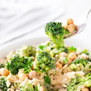 Nahaufnahme einer veganen Brokkoli-Reis-Pfanne mit Kichererbsen auf einer Gabel, im Hintergrund der restliche Pfanneninhalt mit frischer Petersilie.