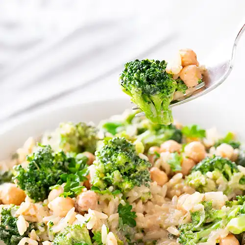 Nahaufnahme einer veganen Brokkoli-Reis-Pfanne mit Kichererbsen auf einer Gabel, im Hintergrund der restliche Pfanneninhalt mit frischer Petersilie.