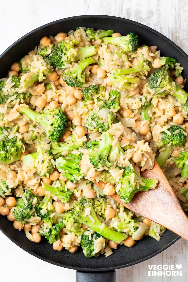 Cremige vegane Brokkoli-Reis-Pfanne mit Kichererbsen in einer schwarzen Pfanne, angerichtet mit einem Holzlöffel.