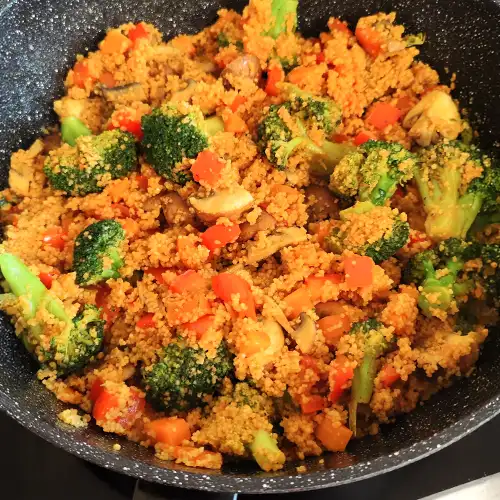 Couscous mit buntem Gemüse, darunter Brokkoli, Paprika und Pilze, frisch gekocht in einer schwarzen Pfanne.