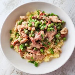 Weißer Teller mit cremiger veganer Hähnchenpfanne mit grünen Erbsen und getrockneten Tomaten, serviert auf Couscous und bestreut mit frischer Petersilie.