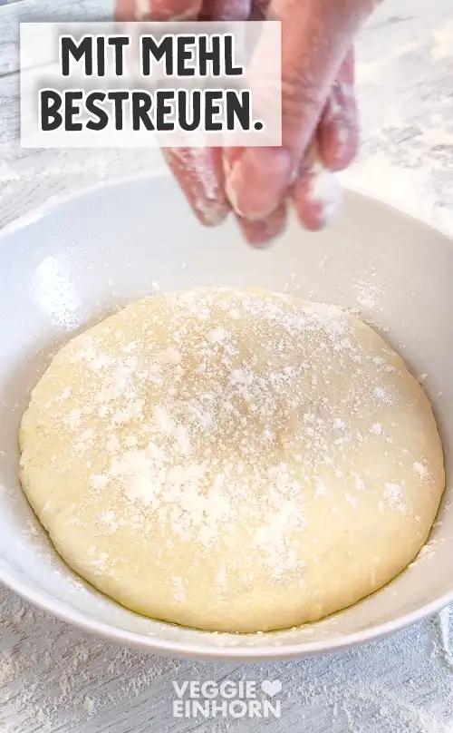 Eine Schüssel mit aufgegangenem Pizzateig, der mit Mehl bestreut wird, eine Hand lässt Mehl über den Teig rieseln.