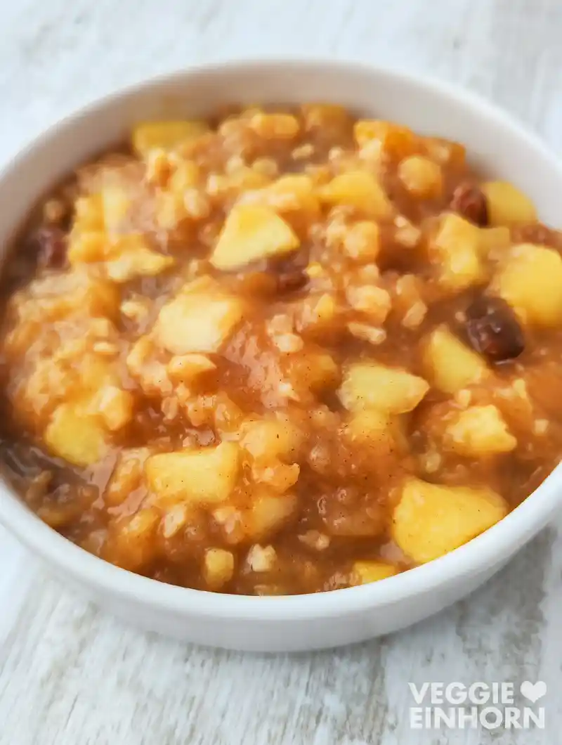 Selbstgemachte vegane Bratapfelsoße mit Apfelstückchen, gehackten Mandeln und Rosinen in einer weißen Schüssel auf hellem Tisch.