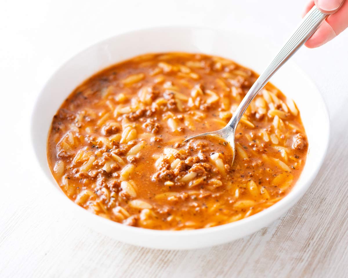 Eine weiße Schüssel mit rötlich-brauner Kritharaki-Suppe, in der kleine Orzo-Nudeln und veganes Hack zu sehen sind. Eine Hand hält einen Löffel, der in die Suppe eingetaucht ist.