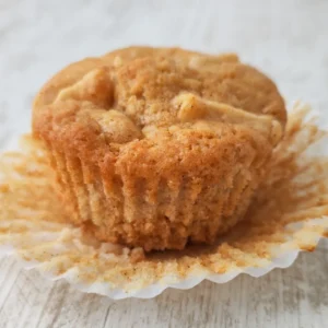 Ein goldbrauner veganer Apfel Muffin liegt im aufgefalteten Papierförmchen auf hellem Holz.