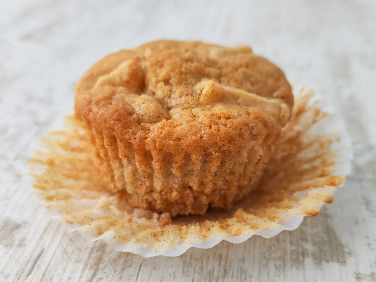 Ein goldbrauner veganer Apfel Muffin liegt im aufgefalteten Papierförmchen auf hellem Holz.