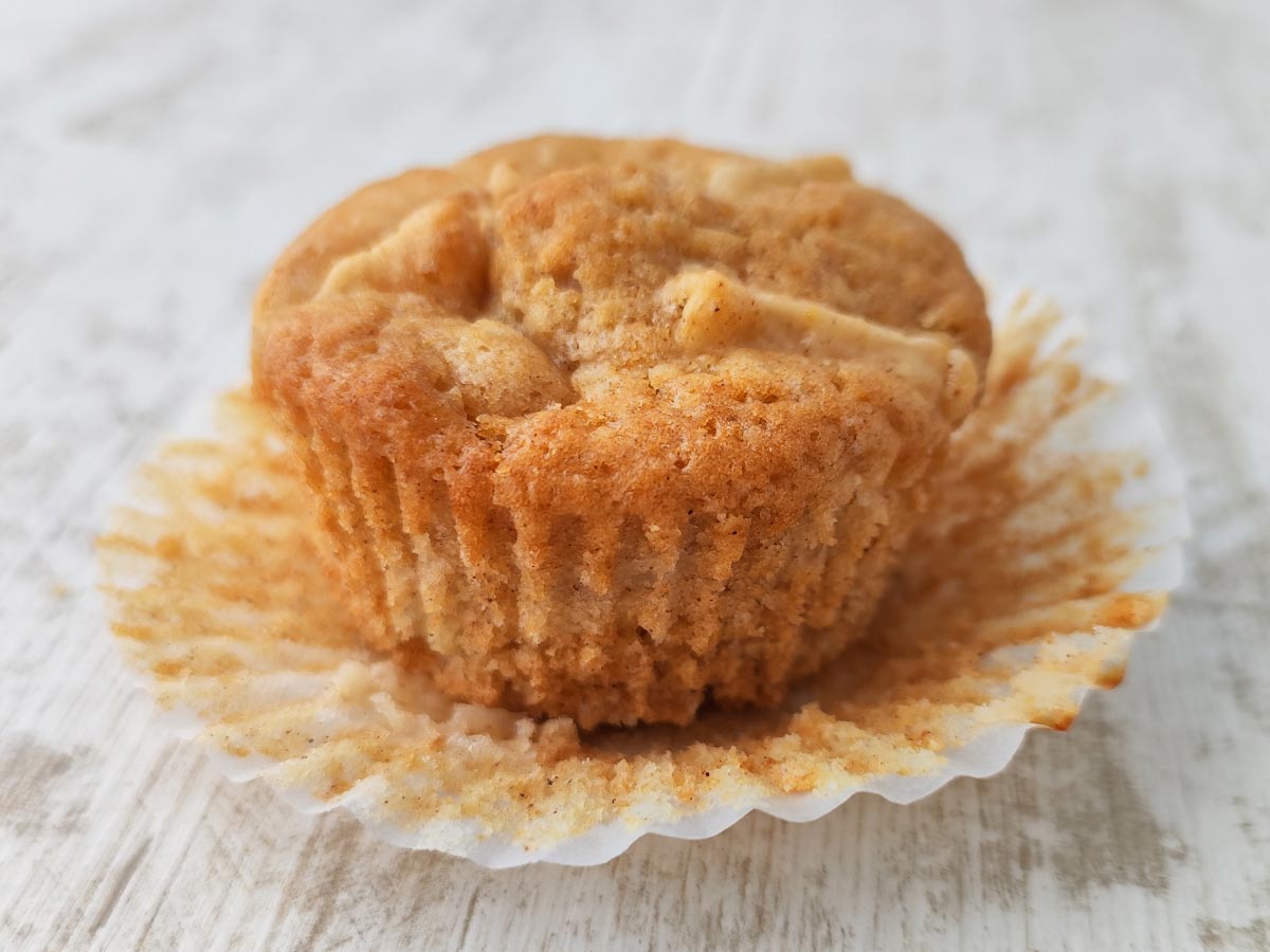Ein frisch gebackener veganer Apfelmuffin im geöffneten Papierförmchen auf hellem Untergrund.