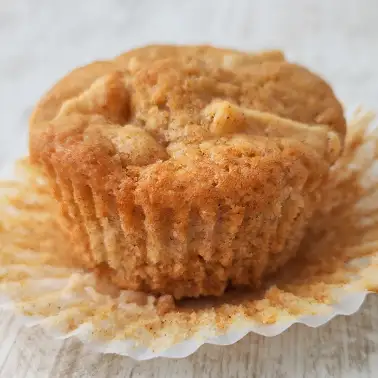 Ein frisch gebackener veganer Apfelmuffin im geöffneten Papierförmchen auf hellem Untergrund.