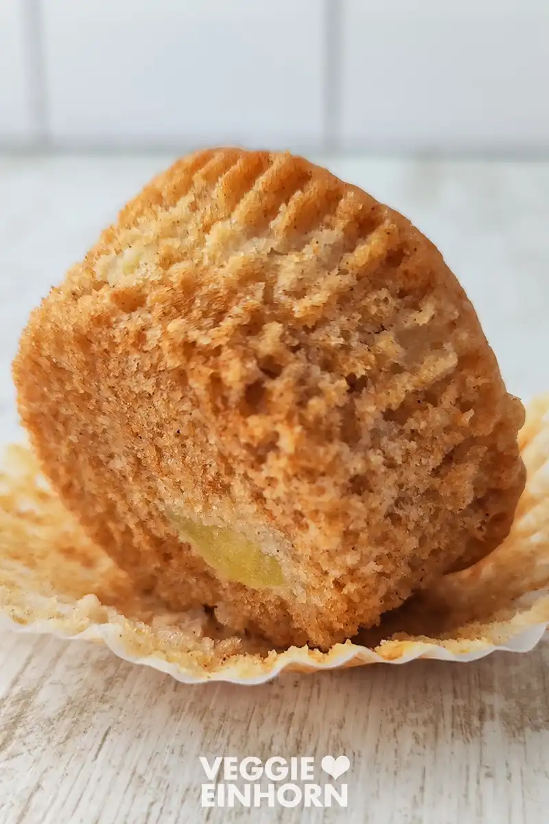 Ein veganer Apfelmuffin auf einem Papierförmchen, seitlich aufgestellt, sodass die saftige und fluffige Krume sichtbar ist.
