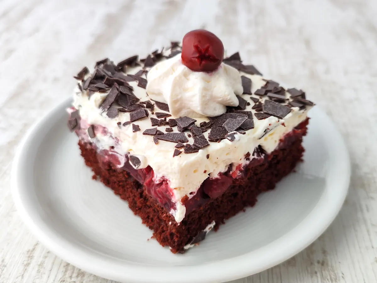 Ein Stück vegane Schwarzwälder Kirschtorte vom Blech auf einem weißen Teller mit Schokoraspeln, Sahne und einer Kirsche als Topping.