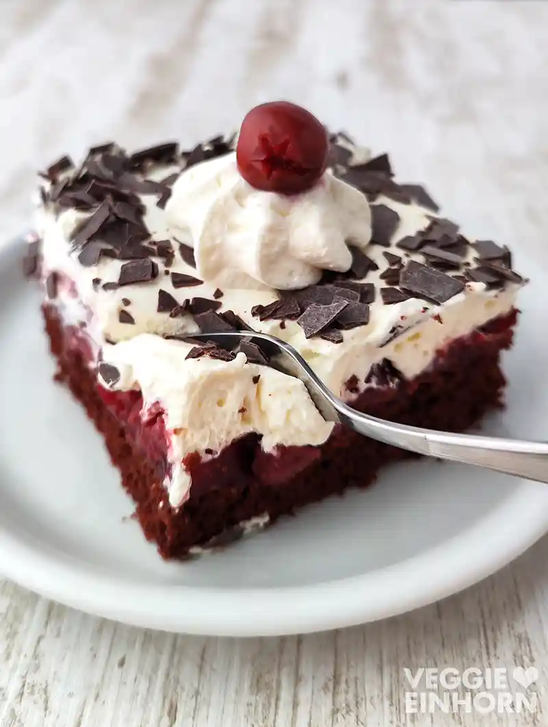 Vegane Schwarzwälder Kirschtorte vom Blech mit Schokoboden, Kirschschicht und Sahne auf einem Teller, eine Gabel nimmt ein Stückchen davon ab.