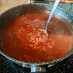 Eine dunkle vegane Zwiebelsoße in einer Pfanne auf dem Herd, mit Zwiebelringen und einer Kelle.