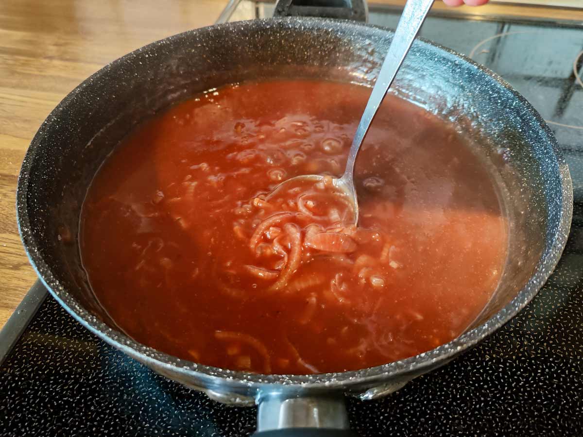 Vegane braune Zwiebelsoße in einer Pfanne auf dem Herd, mit Zwiebelringen und einer Schöpfkelle.