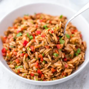 Ein weißer Teller mit Kritharaki Salat mit veganem Hackfleisch, roten Paprikastücken und frischen Kräutern, serviert mit einer Gabel.