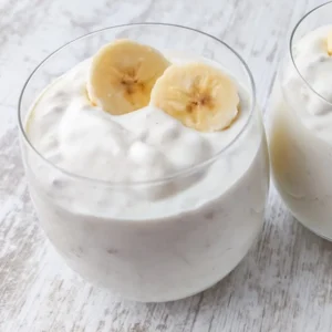 Zwei Gläser mit cremigem veganem Bananenjoghurt, jeweils mit zwei frischen Bananenscheiben garniert, auf hellem Holztisch.