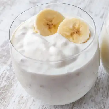 Glas mit cremigem veganem Bananenjoghurt, dekoriert mit zwei frischen Bananenscheiben, auf einem hellem Holztisch.