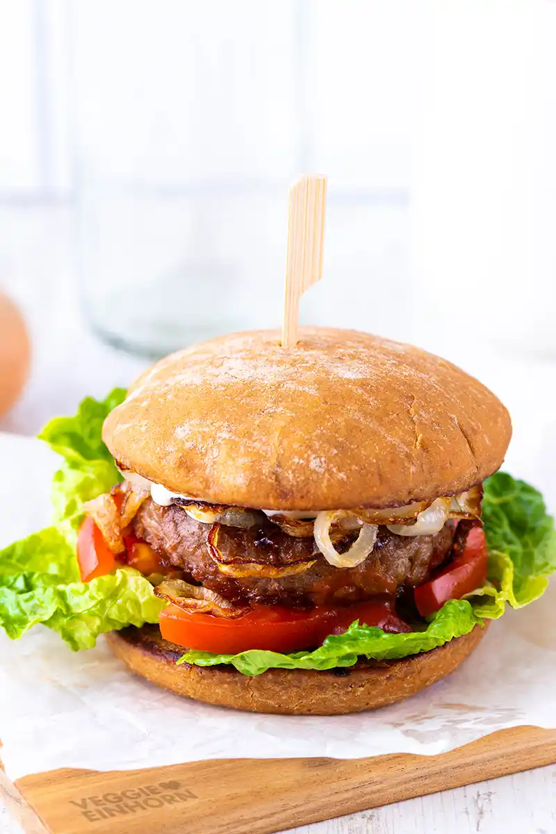 Veganer Burger mit Salat, Tomate, Patty und gebratenen Zwiebeln auf einem hellen Hintergrund mit Holzbrett.