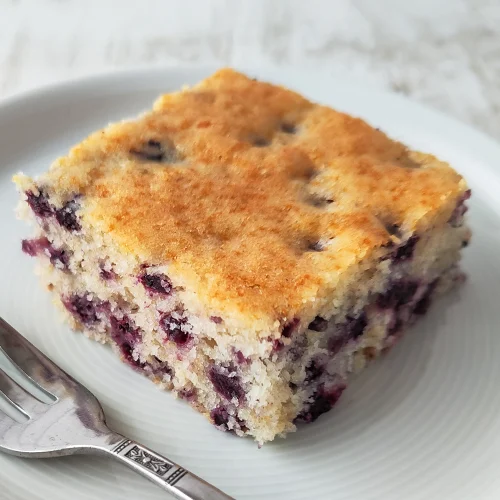Ein Stück veganer Heidelbeerkuchen vom Blech auf einem Teller, daneben eine Kuchengabel.