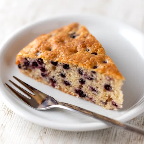 Veganes Kuchenstück mit Heidelbeeren auf einem weißen Teller mit Gabel.