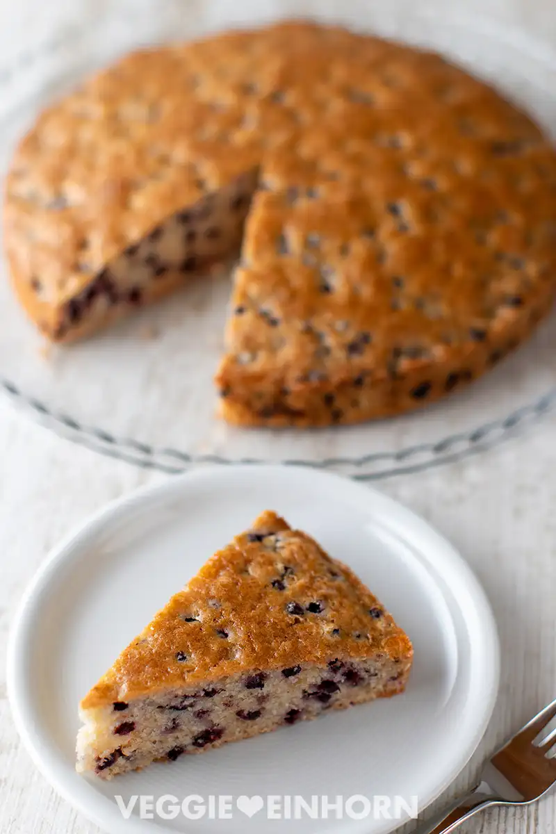 Kuchenstück mit Heidelbeeren auf einem Teller, im Hintergrund der angeschnittene vegane Heidelbeerkuchen auf einer Kuchenplatte.