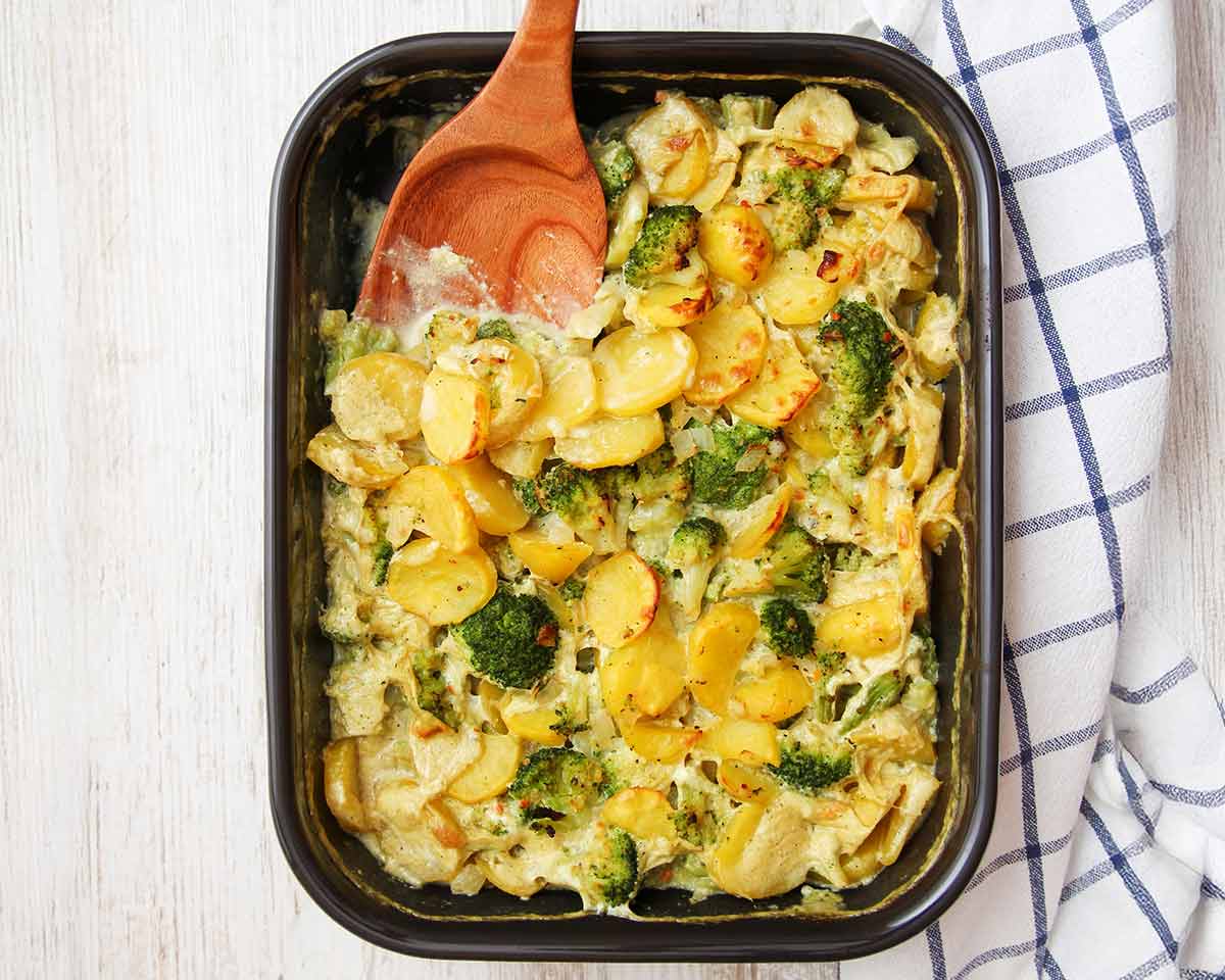 Veganer Kartoffel-Brokkoli-Auflauf in einer schwarzen Form, ein Holzlöffel liegt darin.