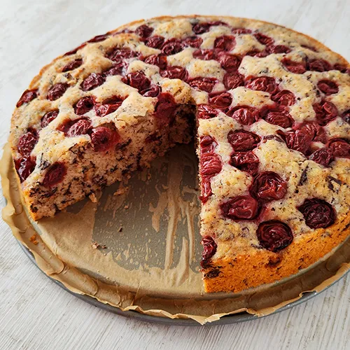 Runder veganer Nusskuchen mit Kirschen und Schokolade, ein Stück ist herausgeschnitten.