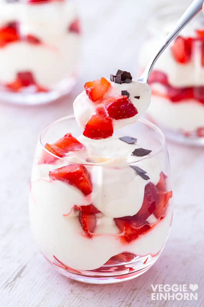 Löffel mit veganem Erdbeer-Dessert über einem Dessertglas, gefüllt mit Erdbeeren, veganer Creme und Schokoraspeln.