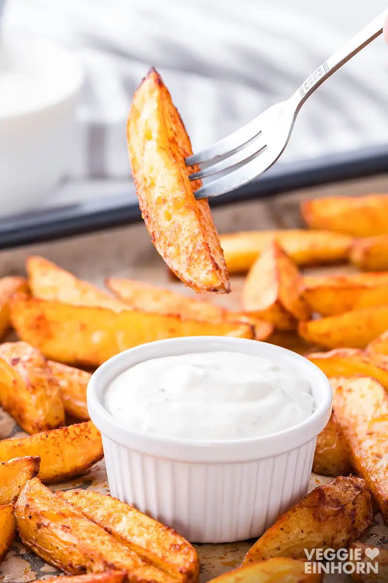 Knusprige Kartoffel Wedges aus dem Ofen, eine Wedge auf einer Gabel wird in eine Schale mit veganem Kräuterquark gedippt.
