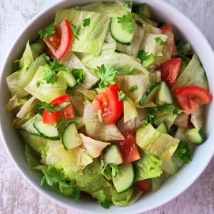 Eisbergsalat mit Tomaten, Gurken und Petersilie in einer weißen Schüssel auf einem hellen Holztisch.