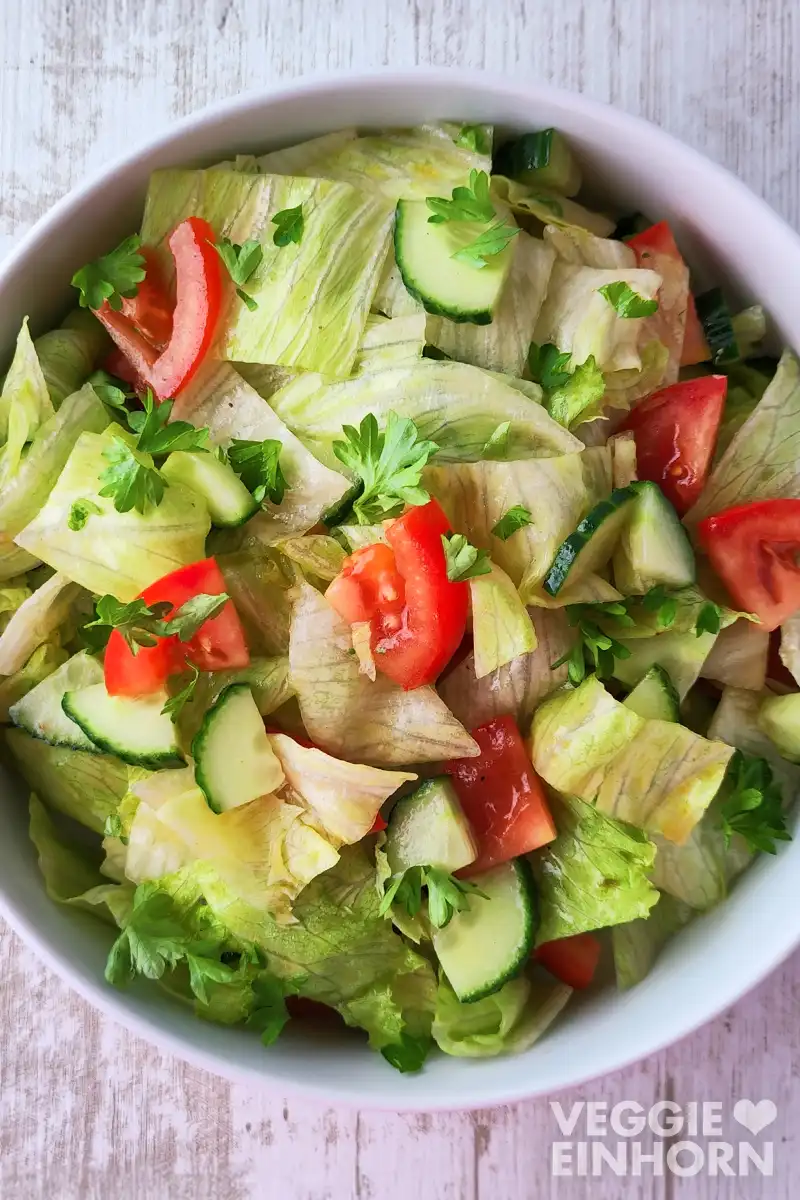 Ein weißer Teller mit Eisbergsalat, Salatgurke, Tomatenstücken und frischer Petersilie auf einem hellen Holztisch.
