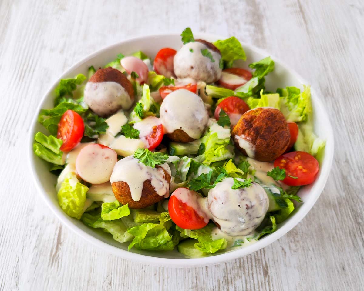 Falafel-Salat mit knusprigen Falafeln, Romanasalat, Kirschtomaten, Gurken und cremigem Dressing, serviert in einer weißen Schüssel auf einem hellen Holztisch.