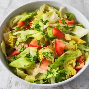 Weiße Schüssel mit frischem Eisbergsalat, Tomatenstücken und gehackter Petersilie auf hellem Untergrund.
