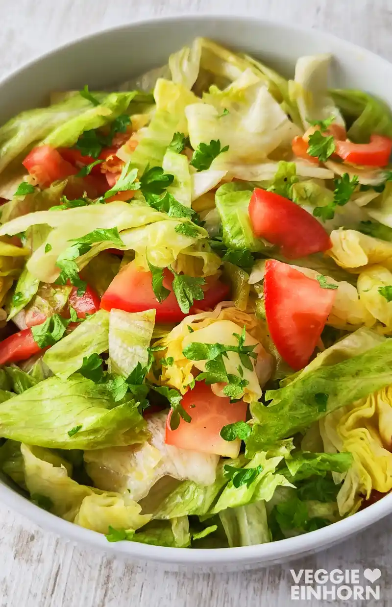 Nahaufnahme eines frischen Eisbergsalats mit Tomatenstücken und gehackter Petersilie in einer weißen Schüssel auf hellem Untergrund.