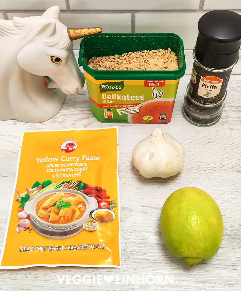 Auf einer hellen Arbeitsfläche stehen geöffnete Packung Gemüsebrühe, eine Pfeffermühle, eine gelbe Currypaste, eine Knoblauchknolle und eine Zitrone neben einer weißen Einhornfigur.
