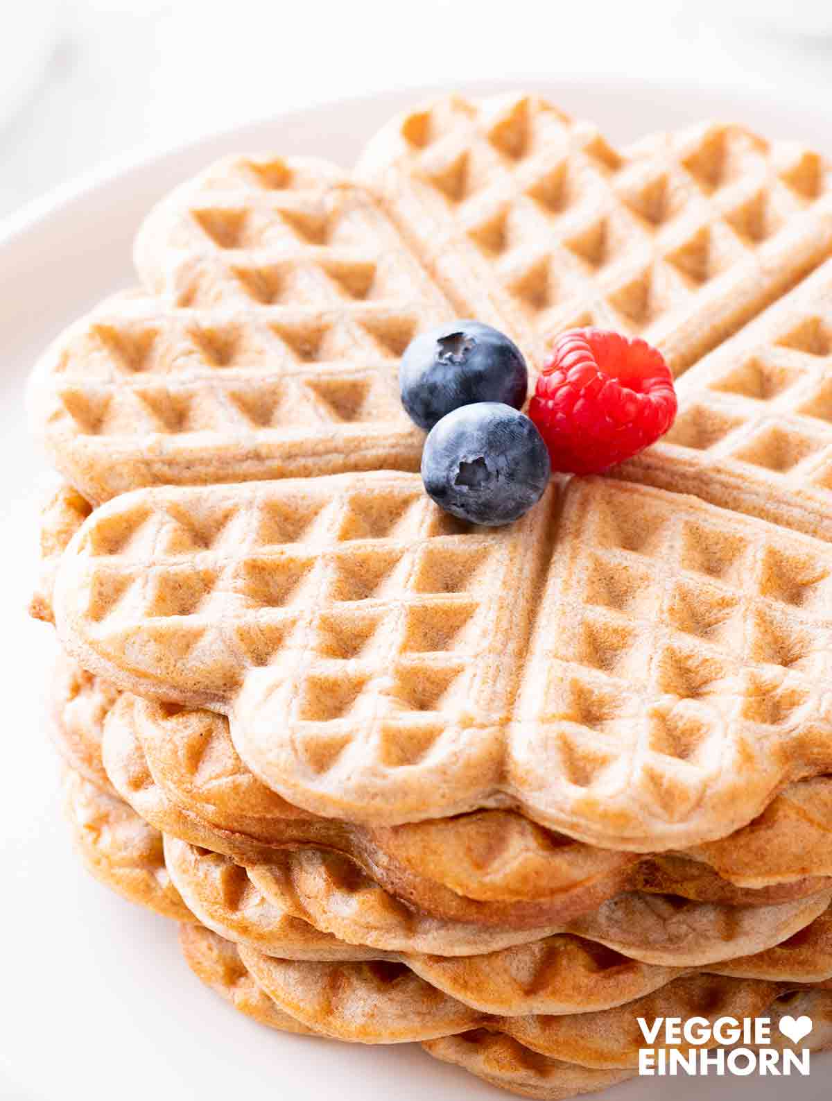 Nahaufnahme eines Stapels herzförmiger veganer Waffeln ohne Zucker, dekoriert mit einer Himbeere und zwei Heidelbeeren.