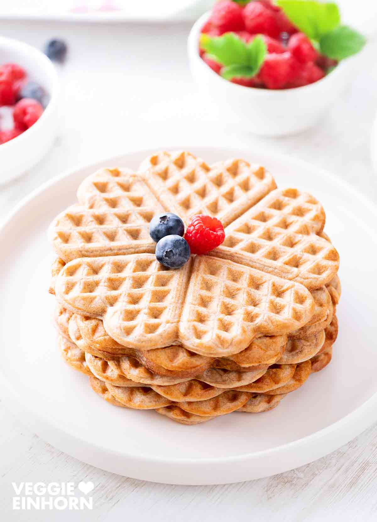 Vegane, zuckerfreie Waffeln in Herzform, gestapelt auf einem weißen Teller, garniert mit frischen Beeren. Im Hintergrund sind Schalen mit Beeren sichtbar.