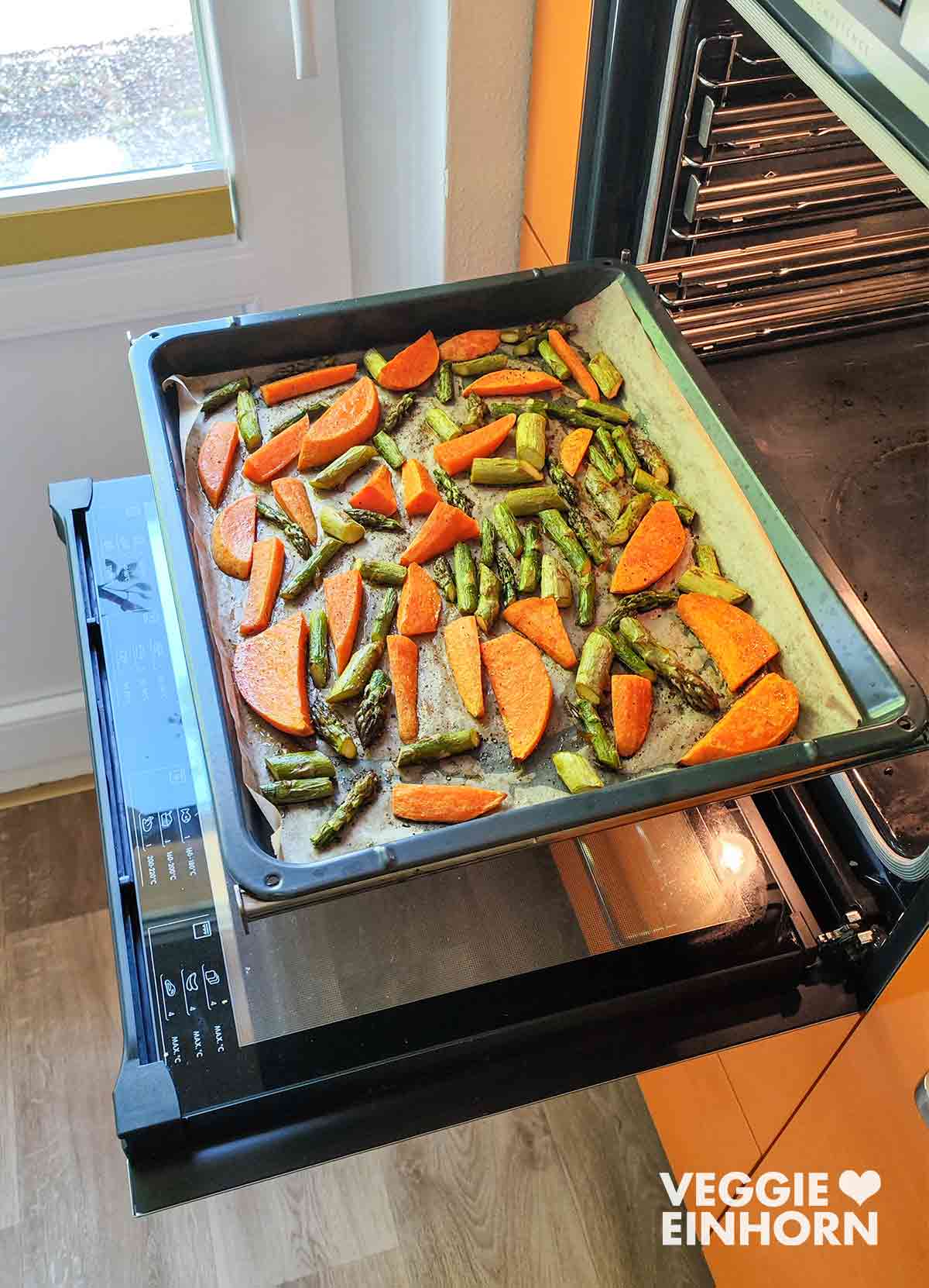 Grüner Spargel und Süßkartoffelspalten im geöffneten Backofen auf einem mit Backpapier belegten Blech.