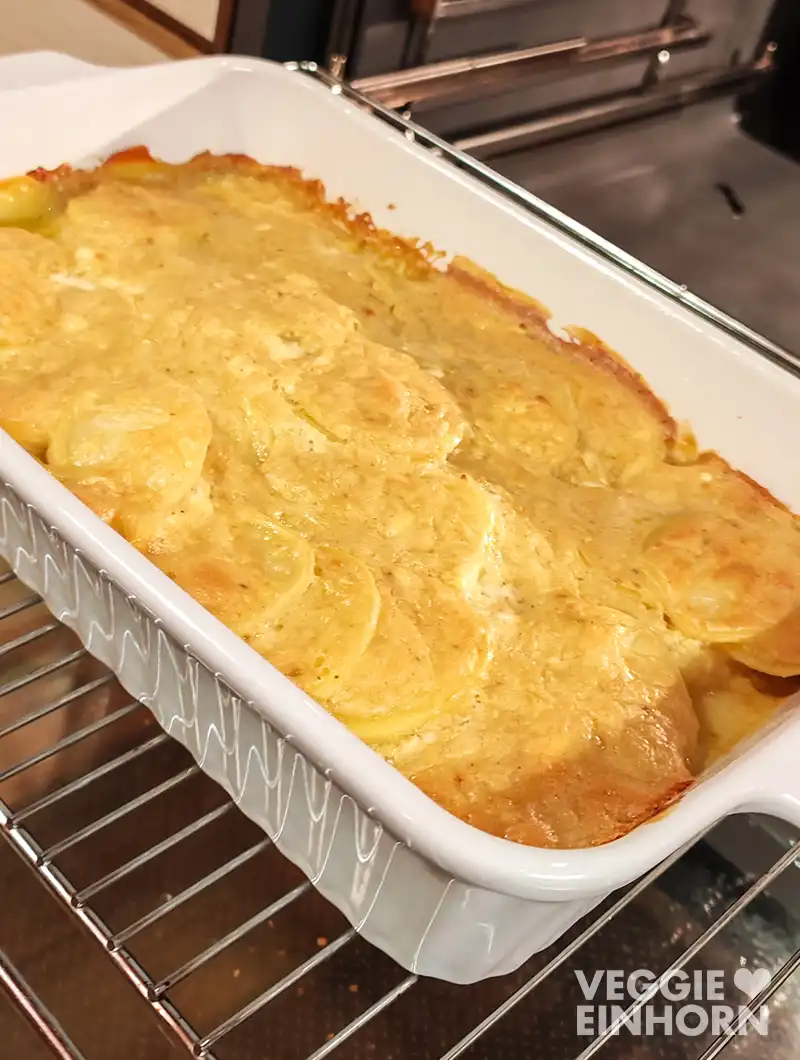 Nahaufnahme einer weißen Auflaufform mit veganem Kartoffelgratin auf dem Ofengitter vor dem geöffneten Ofen.